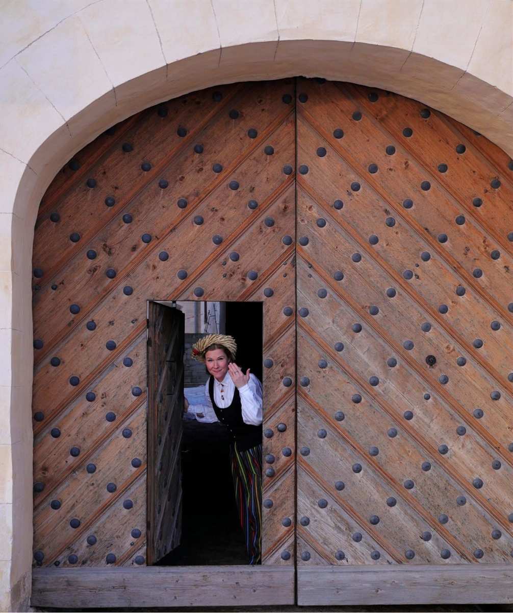 Daiga em traje popular letão tradicional a espreitar pelo portão de madeira cravejado do Palácio de Rundāle