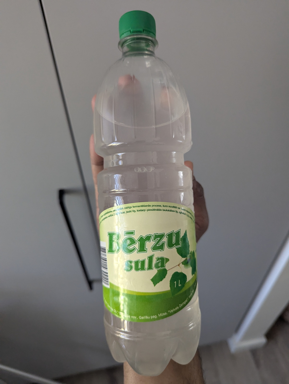 Uma garrafa PET de 1 litro de Bērzu sula (sumo de bétula letão) com um rótulo de padrão de folha verde, levantada contra uma parede de cozinha